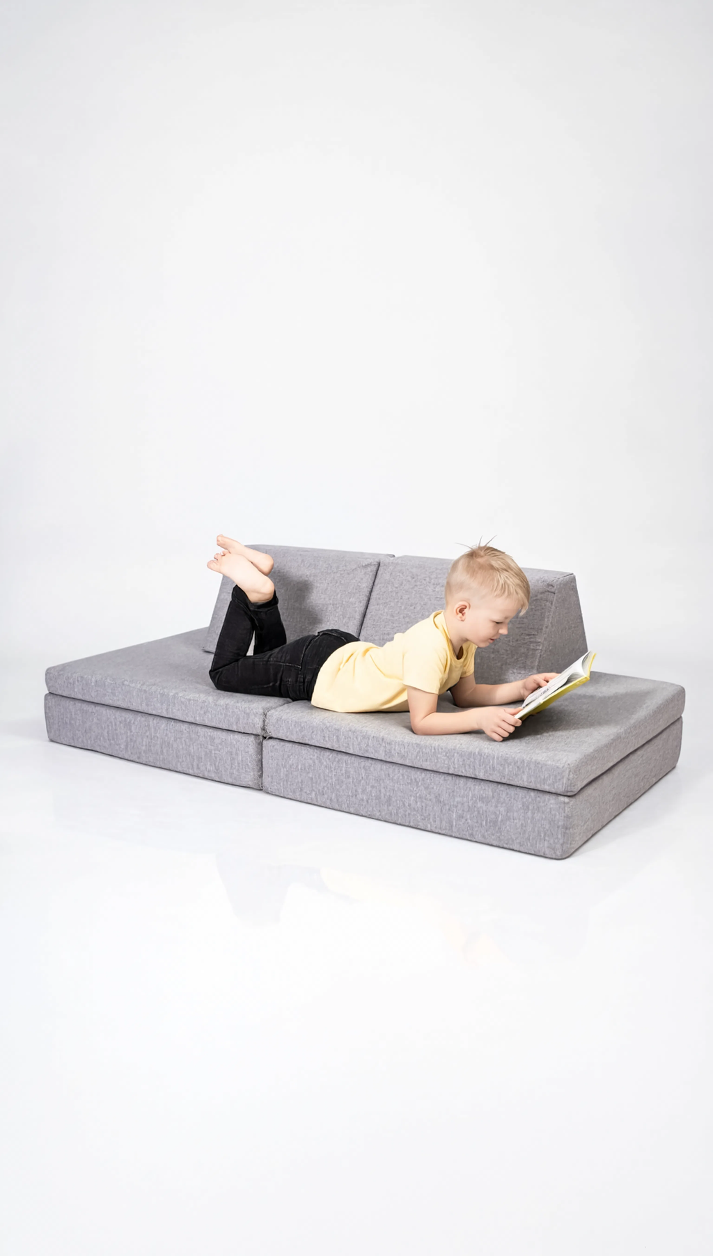 Child lying on a gray sectional sofa reading a book against a white background