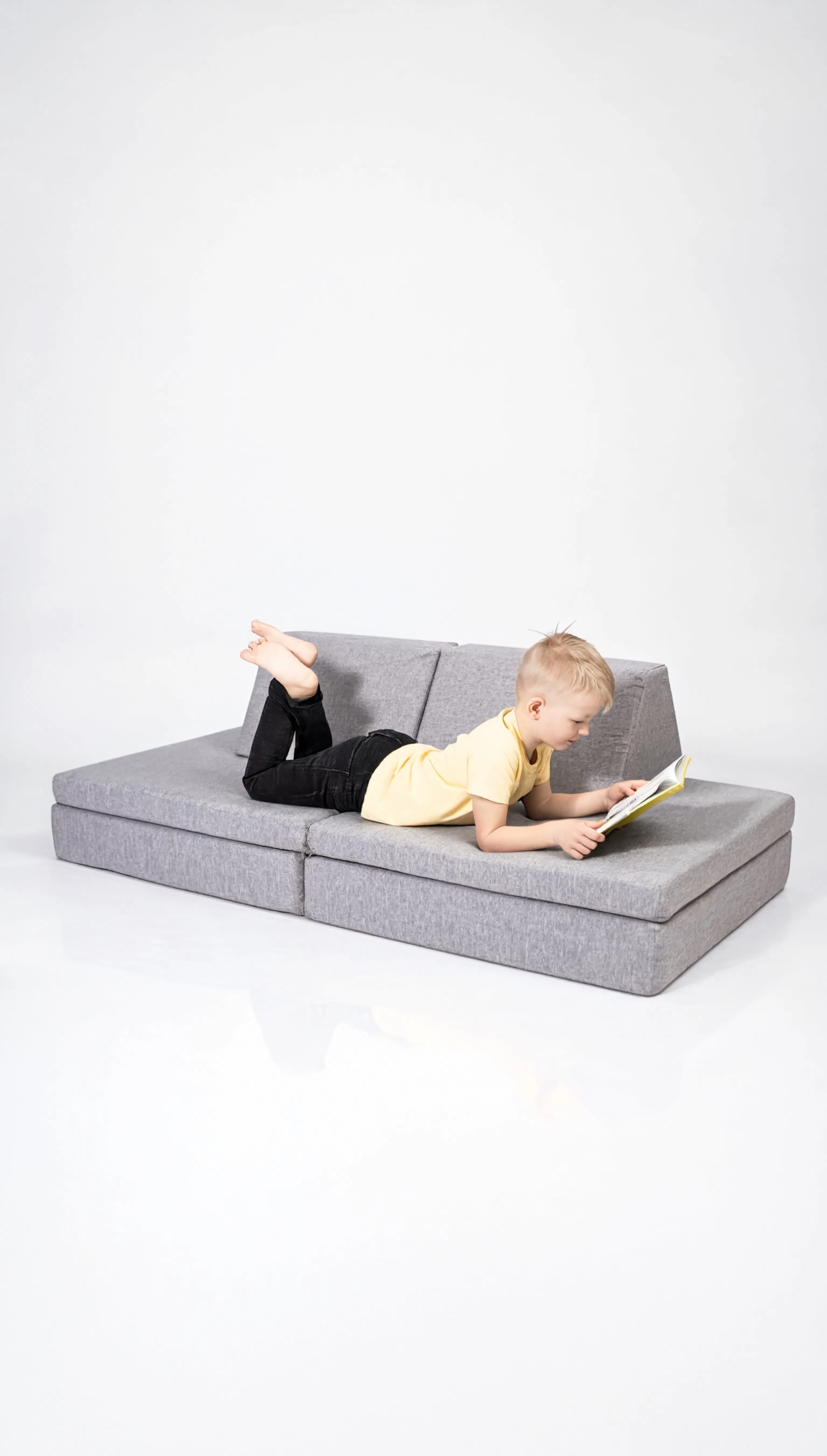 Child lying on a gray sectional sofa reading a book against a white background