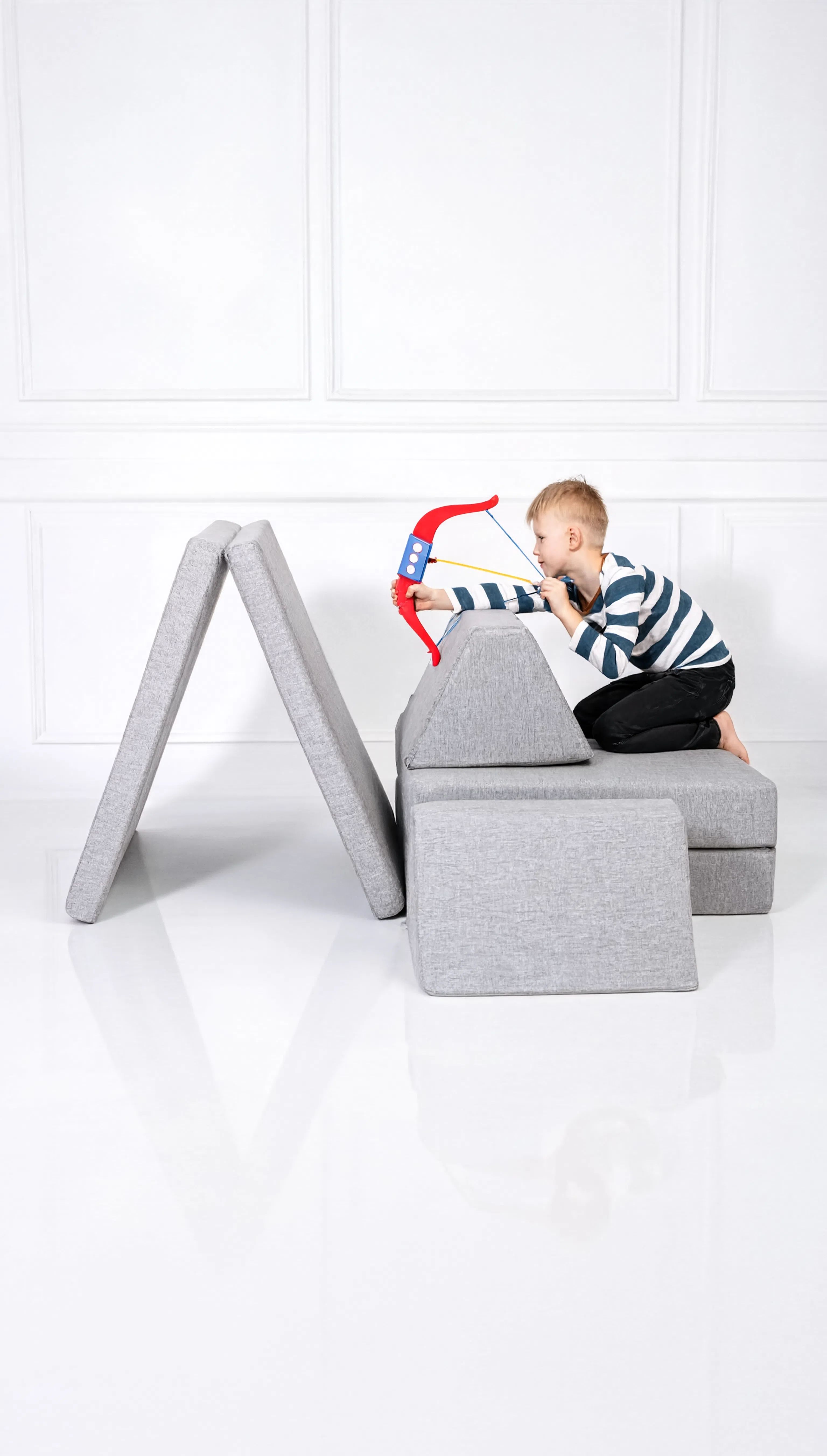 Child playing with a toy bow and arrow on a gray chair against a white background