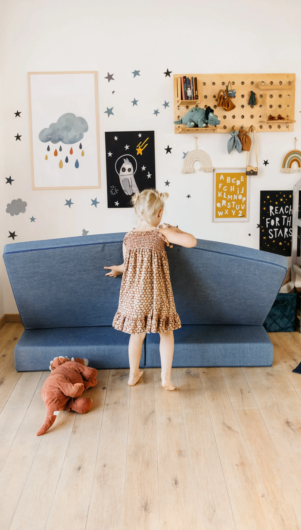Child in a patterned dress standing next to a blue sofa in a room with wall decorations and a teddy bear on the floor.
