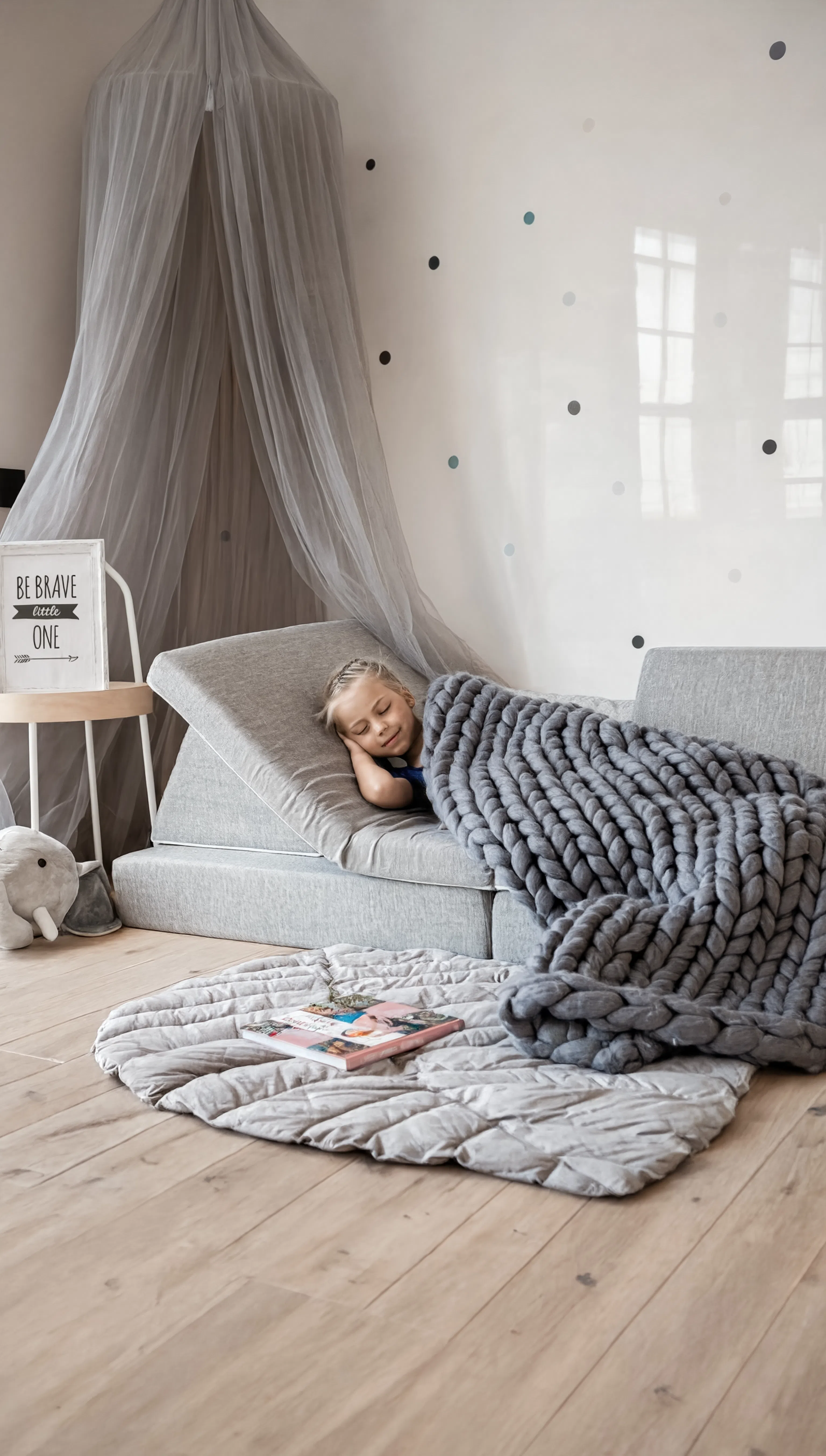 Child lying on a gray sofa under a canopy in a cozy room with a rug and books.