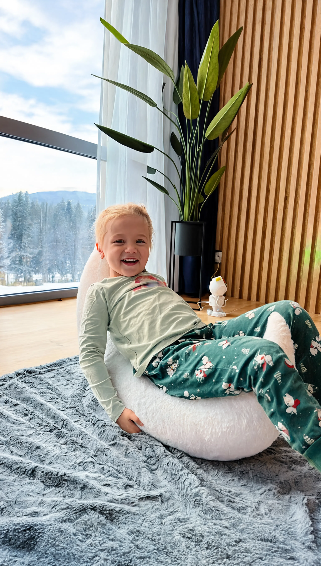 Child in green pajamas sitting on a white cushion by a window with a snowy landscape view.
