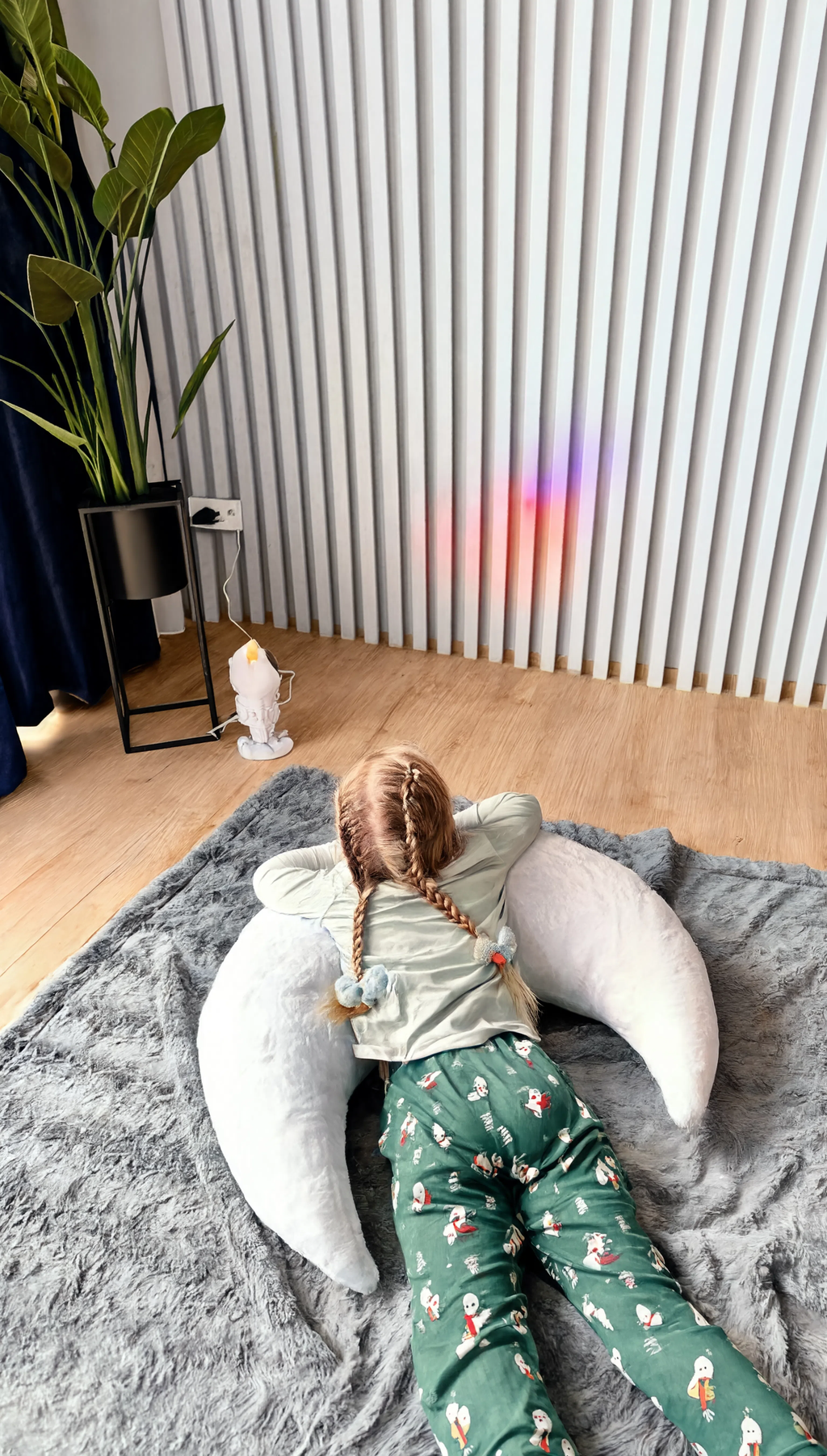Child sitting on a gray rug with a white pillow, wearing green pajama pants with a pattern.