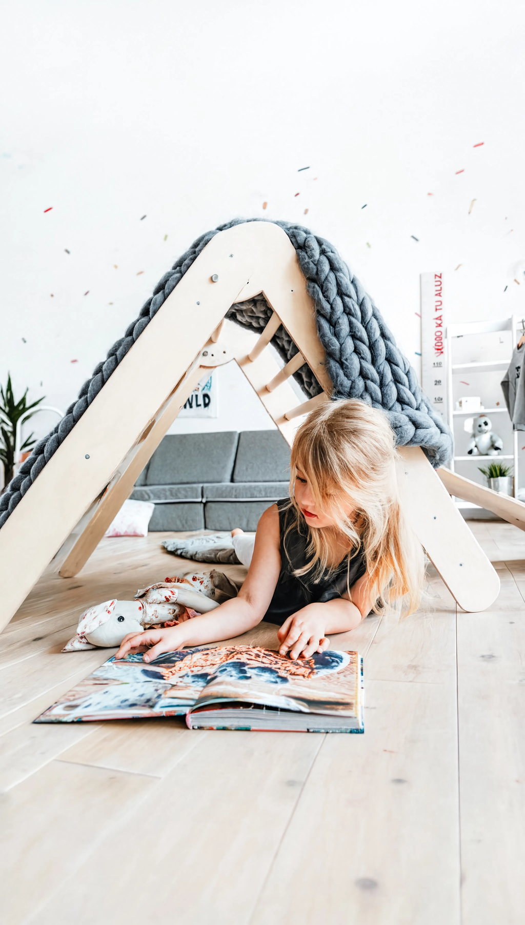 Child reading a book on the floor next to a wooden playhouse with a textured gray cover.