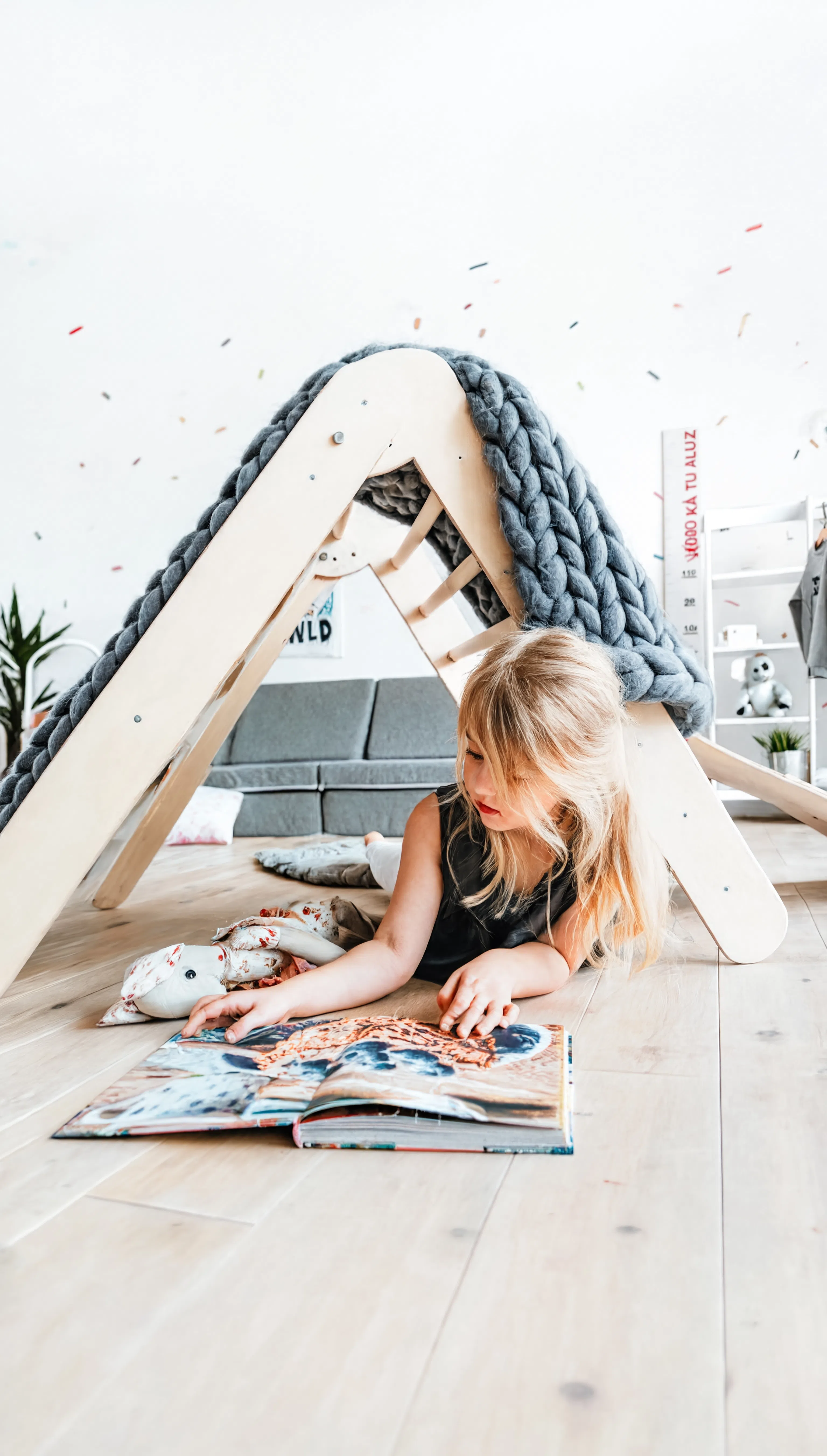 Child reading a book on the floor next to a wooden playhouse with a textured gray cover.