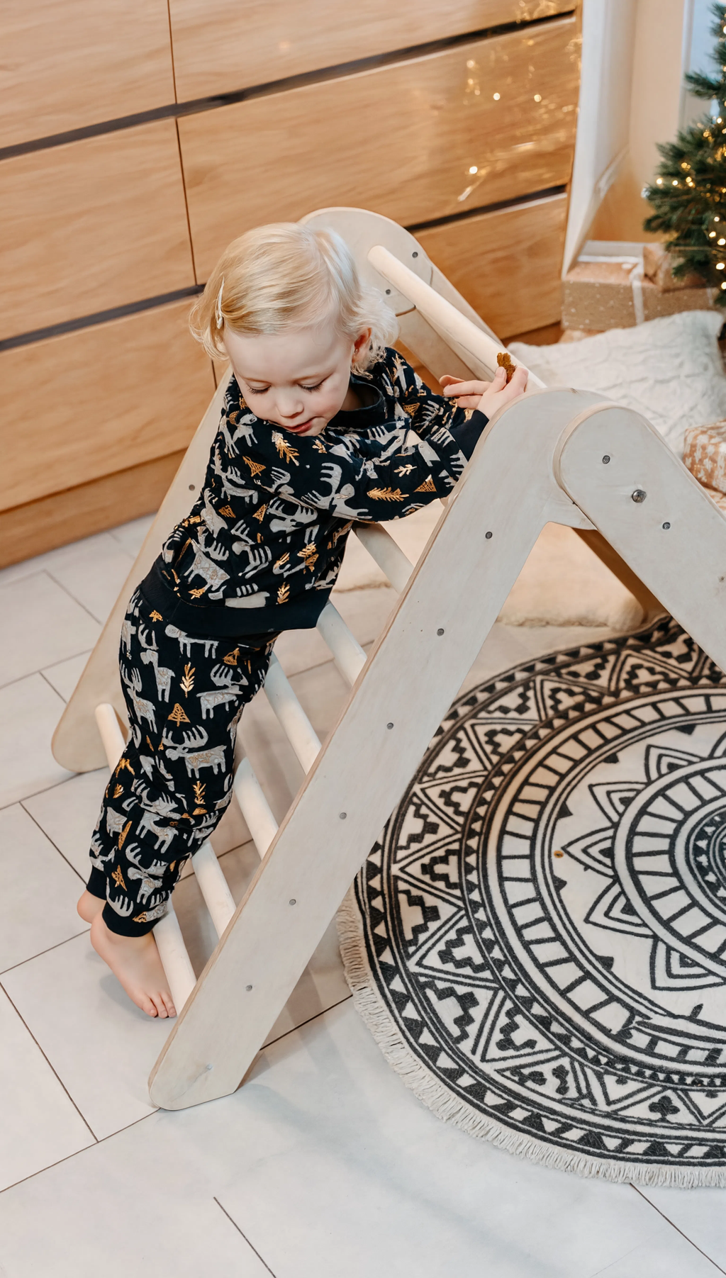Child playing on a wooden climbing toy in a room with a round rug and Christmas tree.