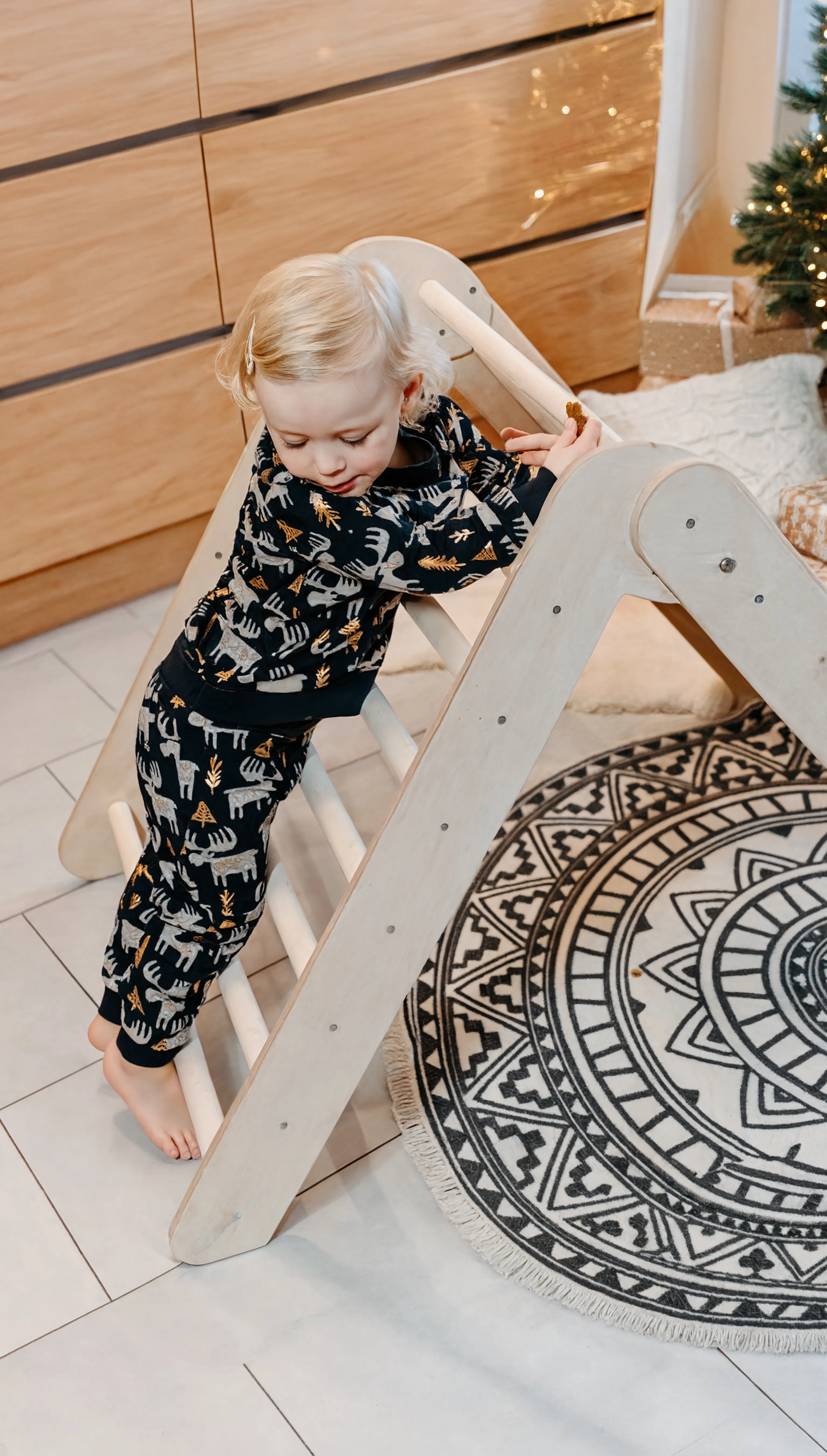 Child playing on a wooden climbing toy in a room with a round rug and Christmas tree.