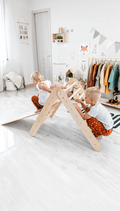 Two children playing on a wooden climbing toy in a room with white walls and minimal decor.