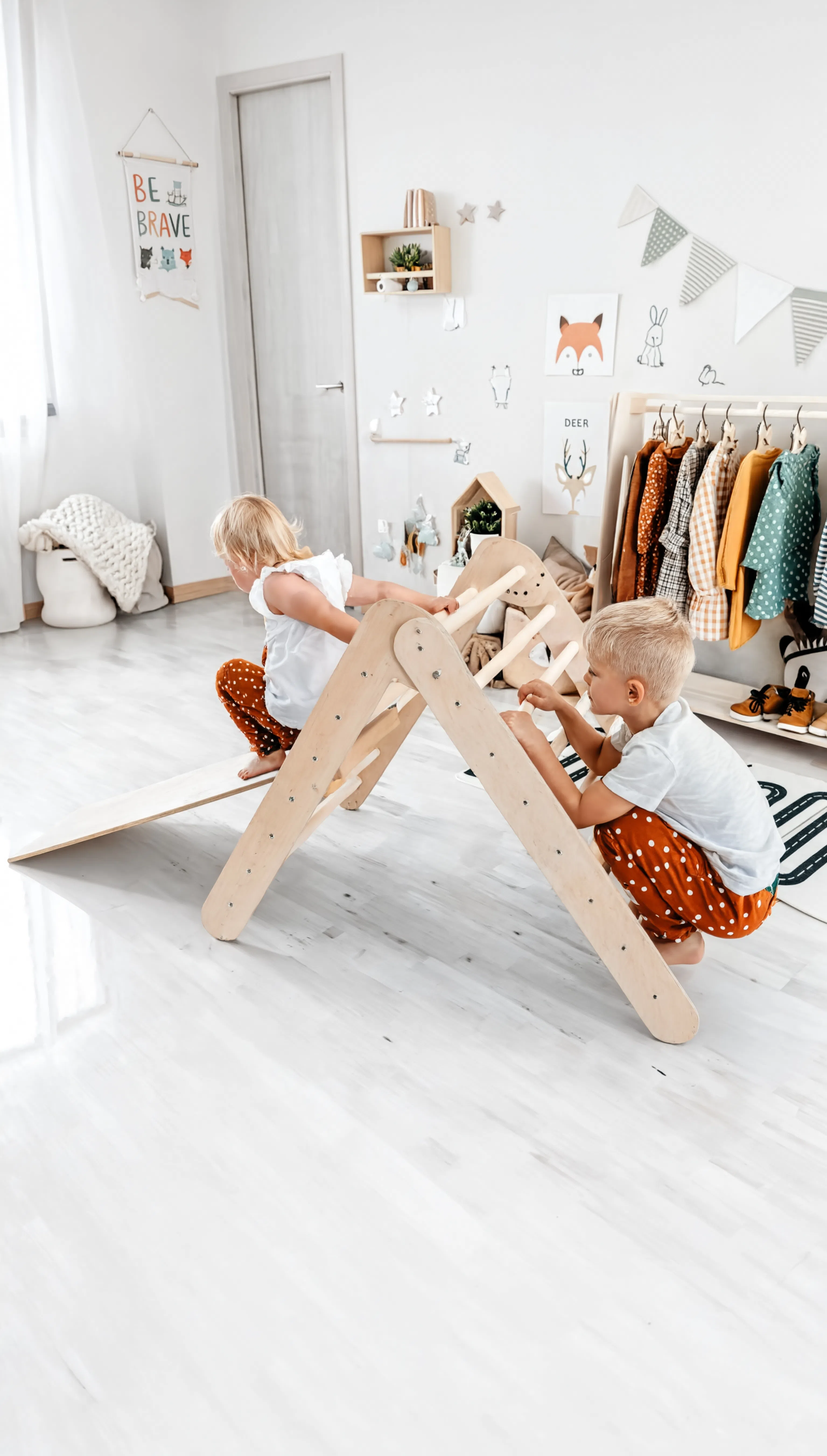 Two children playing on a wooden climbing toy in a room with white walls and minimal decor.