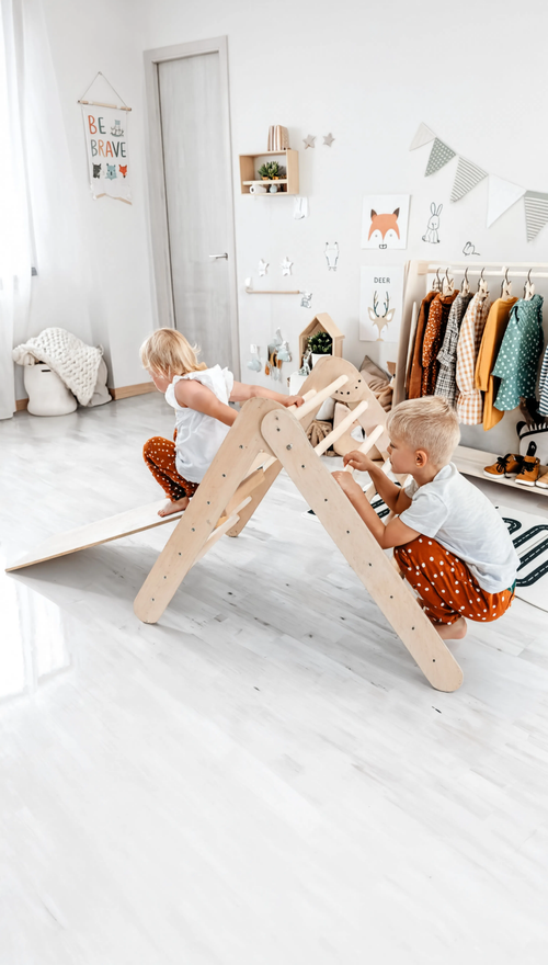 Two children playing on a wooden climbing toy in a room with white walls and minimal decor.