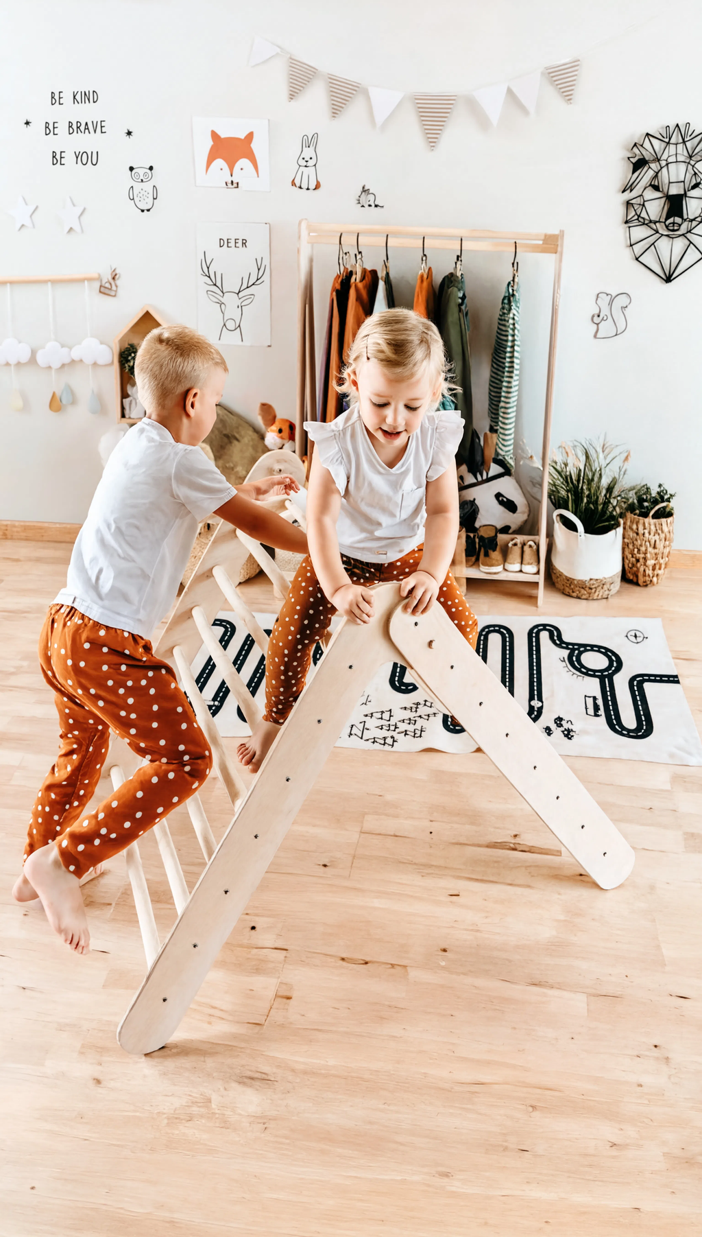Two children playing on a wooden climbing toy in a room with decor elements.