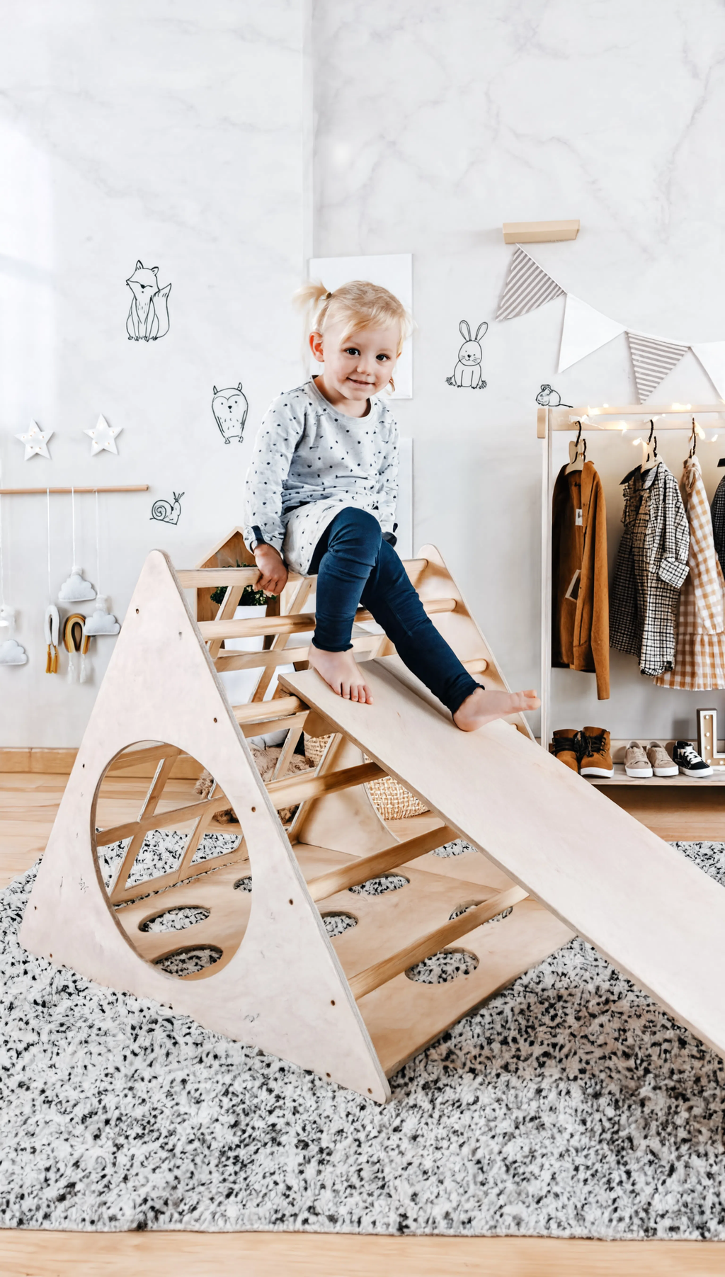 Child playing on a wooden climbing toy in a room with clothes hanging on a rack.