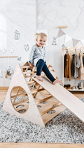 Child playing on a wooden climbing toy in a room with clothes hanging on a rack.
