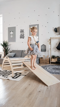 Child standing on a wooden seesaw in a playroom with toys and furniture.
