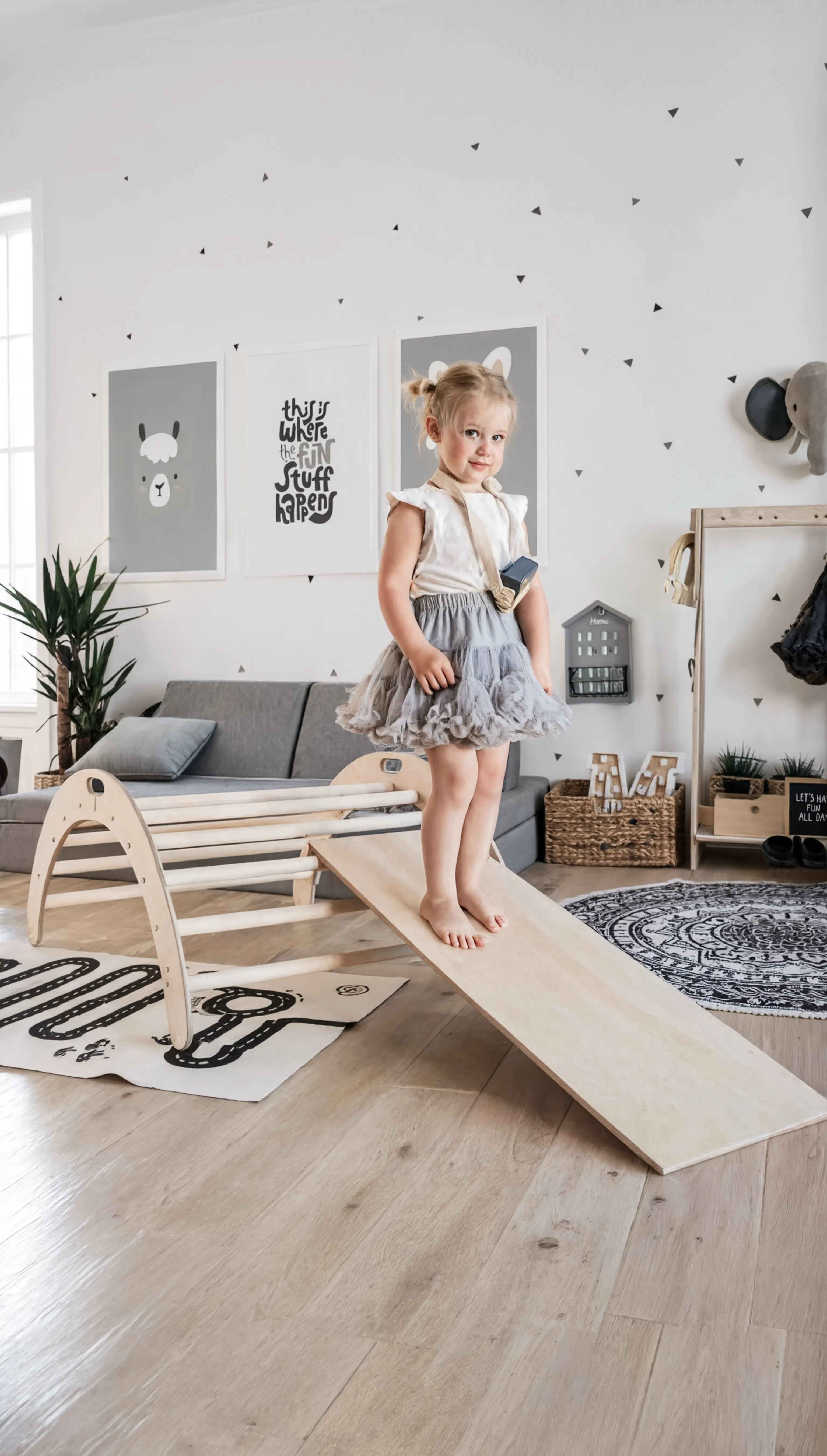 Child standing on a wooden seesaw in a playroom with toys and furniture.