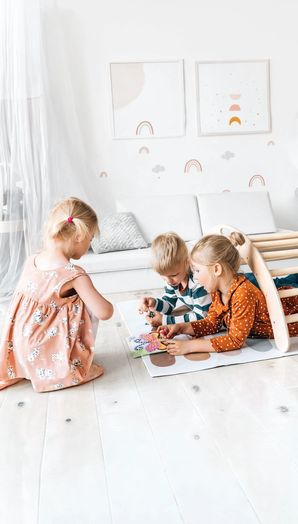 Three children sitting on a white floor in a room with colorful wall art.