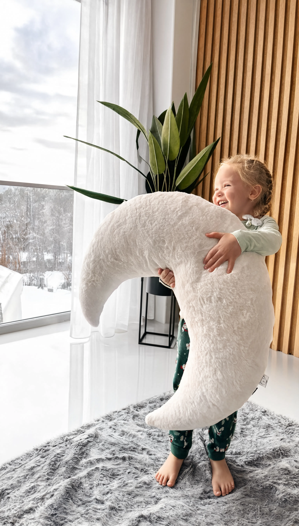 Child holding a large white crescent moon-shaped pillow in a room with a snowy outdoor view.